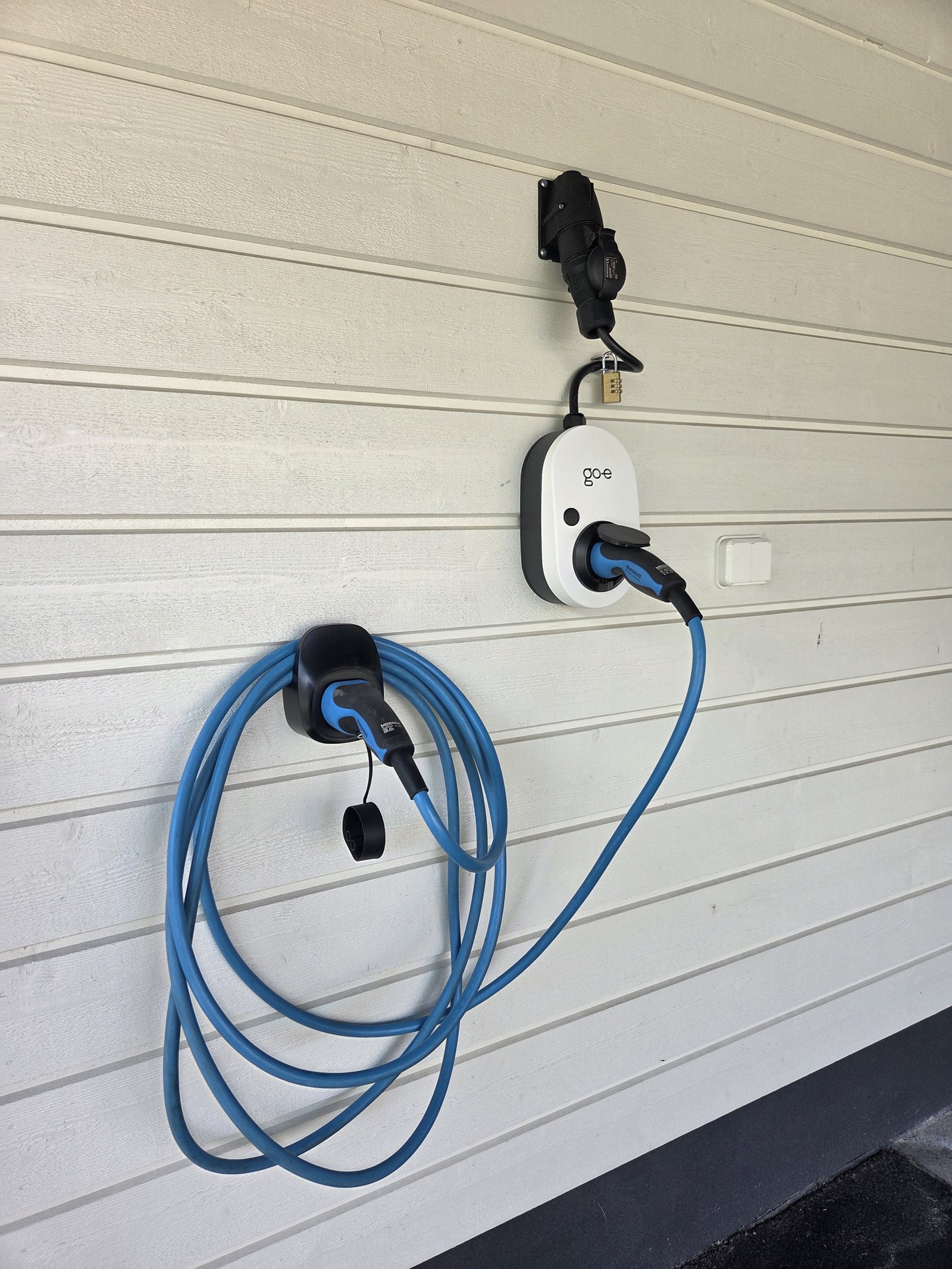 go-e Charger Gemini Flex autokatoksen seinällä: ylhäällä voimavirtarasia, jossa laturi kiinni, ja latauskaapeli siististi pidikkeellään. Pistokkeen läpi kulkee munalukko.