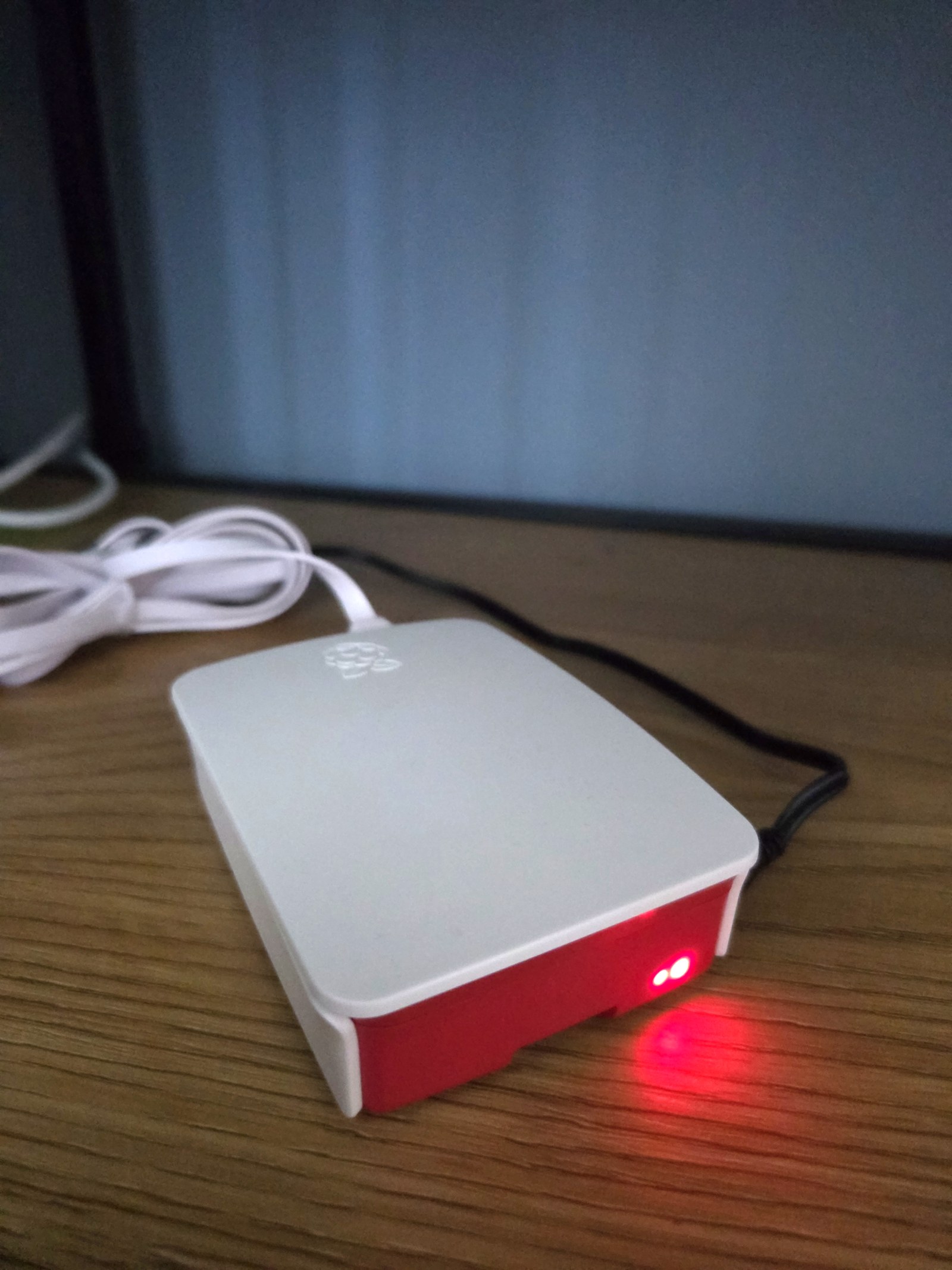 Raspberry Pi 3 virallisessa puna-valkoisessa kotelossaan hyllynkulmassa, punainen merkkivalo hehkuu hämärässä puupintaa vasten.